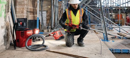 Trabajador en un sitio de construcción, usando herramientas eléctricas y equipado con casco y chaleco.