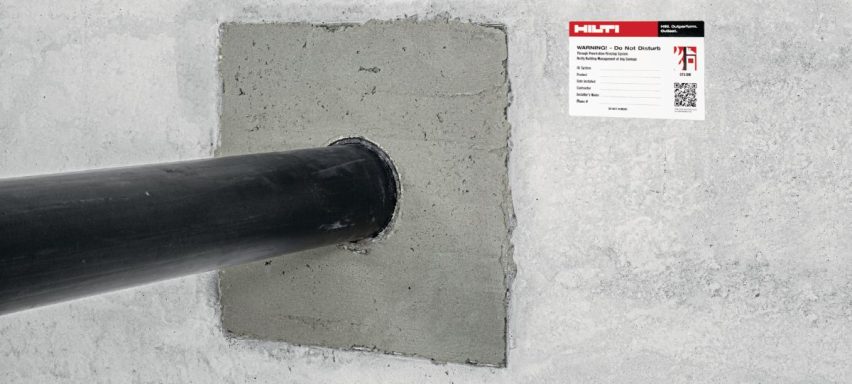 Tubería negra sale de una pared gris con una etiqueta roja y blanca al lado.