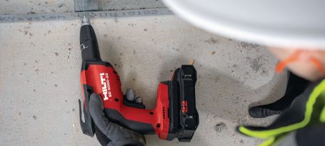 Taladro inalámbrico rojo sobre una superficie de concreto, mano con guantes cerca.