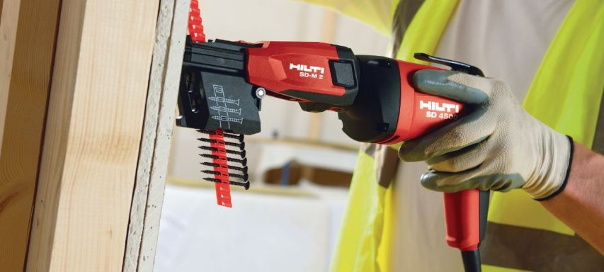 Herramienta eléctrica roja y negra sujetada por una mano con guante, perforando una superficie de madera.