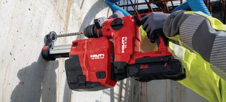 Taladro rotativo rojo siendo utilizado en una pared de concreto. مانوس con guantes.