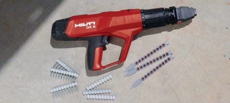 Herramienta eléctrica roja y negra junto a tornillos y clavos en una superficie de concreto.