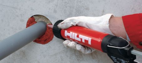 Mano con guante sosteniendo una herramienta Hilti junto a un tubo en una pared.